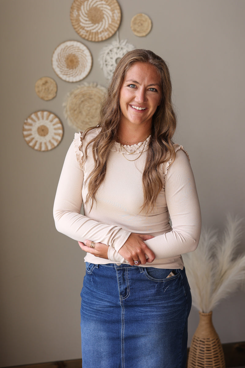 A smiling woman with long light brown hair stands indoors wearing a cream ribbed long-sleeve top with lace ruffle details at the shoulders and a blue denim skirt. She is posed in front of a beige wall decorated with circular woven wall art and a vase of pampas grass.