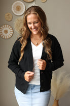 A woman wearing a black button-front cardigan over a white V-neck top and light blue jean skirts, standing indoors against a neutral wall with decorative woven wall art and dried grass in the background.