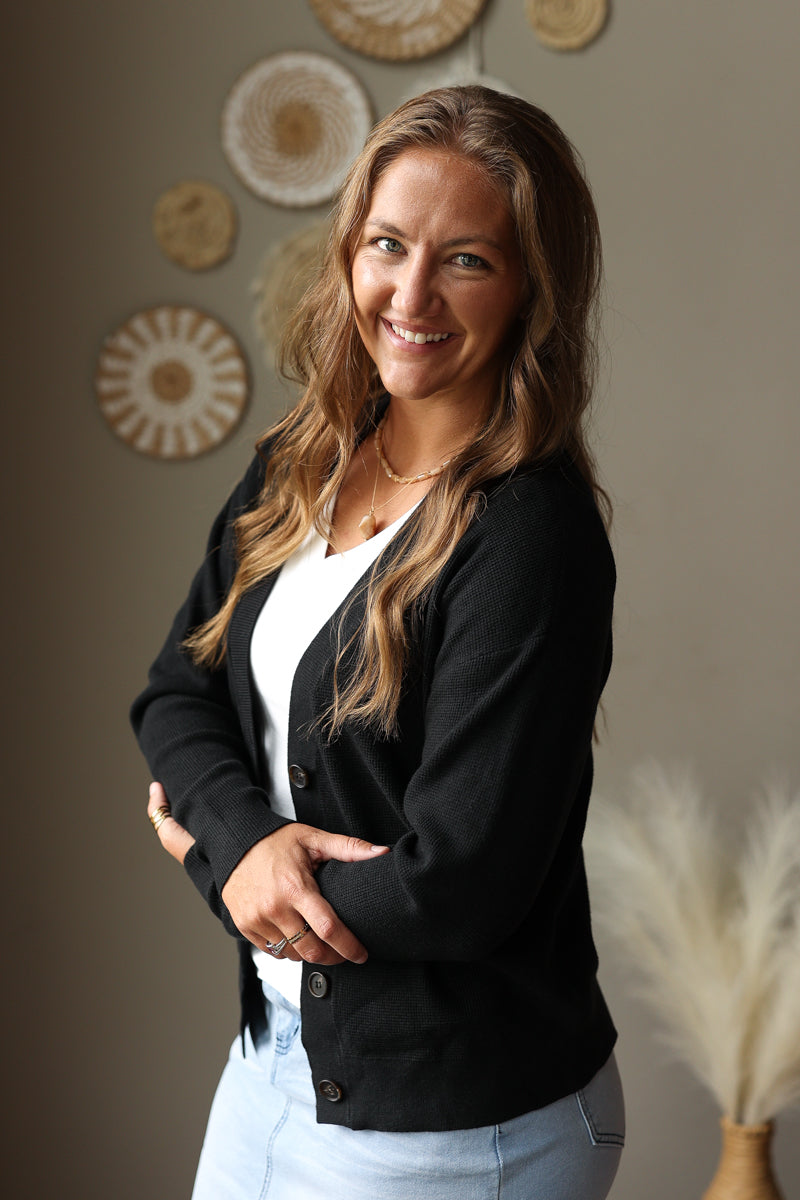 A smiling woman with long light brown hair stands indoors wearing a black button-front cardigan over a white top and light blue jean skirts. The background is a warm neutral wall with decorative woven wall art and dried grass decor.