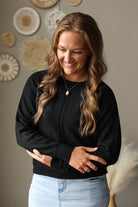 A woman with long wavy light brown hair is wearing a black knit sweater and light blue jean skirt, standing in front of a neutral wall decorated with circular woven wall art.