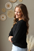 A woman with long brown hair is shown from the waist up, wearing a black knit sweater and light blue jean skirt, standing in front of a neutral wall decorated with woven circular wall art.
