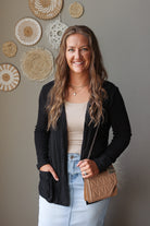 A woman smiling and posing in a black ribbed cardigan over a beige tank top and light blue jean skirt, holding a brown quilted shoulder bag. She is standing against a gray wall decorated with circular woven wall art.
