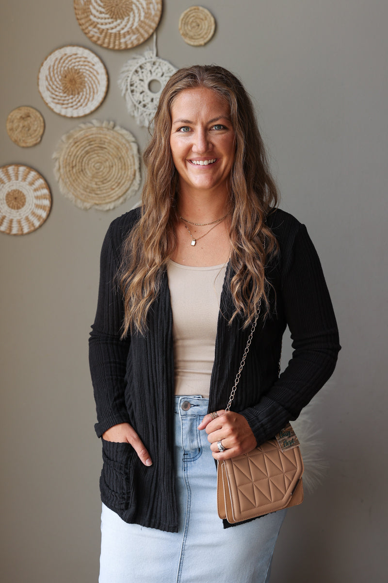 A woman smiling and posing in a black ribbed cardigan over a beige tank top and light blue jean skirt, holding a brown quilted shoulder bag. She is standing against a gray wall decorated with circular woven wall art.