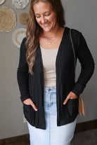 A woman wearing a black ribbed open-front cardigan with pockets over a beige tank top and light blue jean skirt, standing indoors against a gray wall with woven decor.