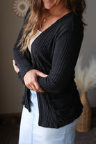 A woman wearing a black ribbed cardigan over a cream top and white skirt, standing indoors with her arms crossed. The background shows a neutral wall with a decorative sunburst and a vase with dried grass.