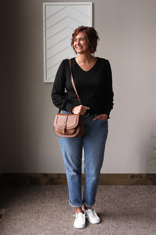 Woman wearing a black top, blue jeans, and white sneakers, holding a brown leather bag indoors.