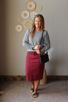 A woman stands indoors wearing a black and white gingham long-sleeve blouse with ruffled shoulders and a high-waisted burgundy midi skirt. She is carrying a black shoulder bag and wearing flat sandals, posed against a neutral wall with decorative woven wall art.