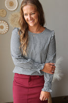 A woman wearing a black and white gingham long-sleeve blouse with ruffled shoulders and a dark red skirt, standing against a neutral wall with decorative woven wall art.