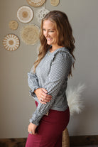 A woman wearing a black and white gingham long-sleeve blouse with ruffled shoulders and burgundy denim skirt, standing in a room with a neutral gray wall decorated with woven wall art.