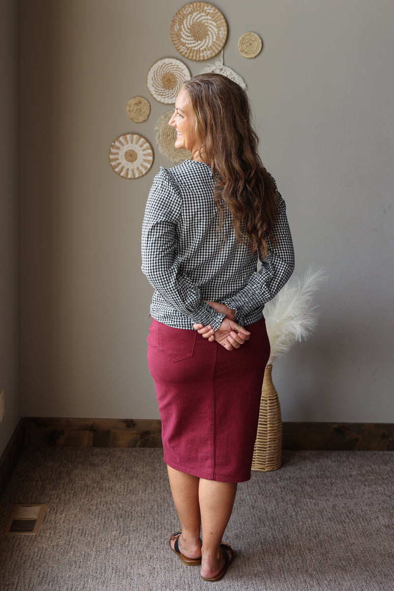 A woman stands in profile wearing a black and white gingham long-sleeve blouse and a burgundy knee-length skirt, standing in a room with a neutral gray wall decorated with circular wall art.