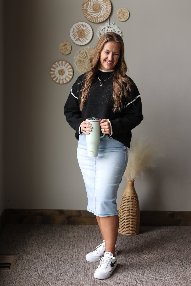 Full outfit view of a black stitch detail sweater with a light denim midi skirt and white sneakers.