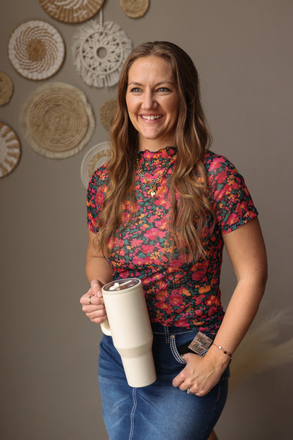 Woman holding a white tumbler with decorative wall in the background, wearing a spring summer black mesh floral top
