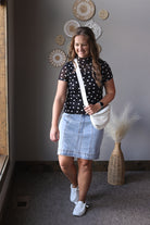 Woman wearing a black polka dot top and denim skirt, holding a white bag, standing indoors.