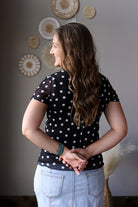 Woman wearing a black polka dot shirt and light blue jean skirt standing in a room with decorative wall art.