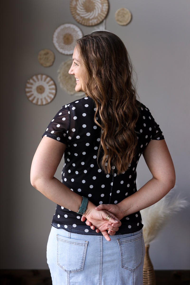 Woman wearing a black polka dot shirt and light blue jean skirt standing in a room with decorative wall art.