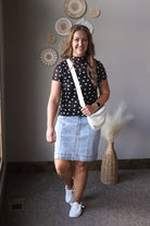 Woman wearing a black polka dot top and denim skirt, standing indoors with decorative wall art in the background.