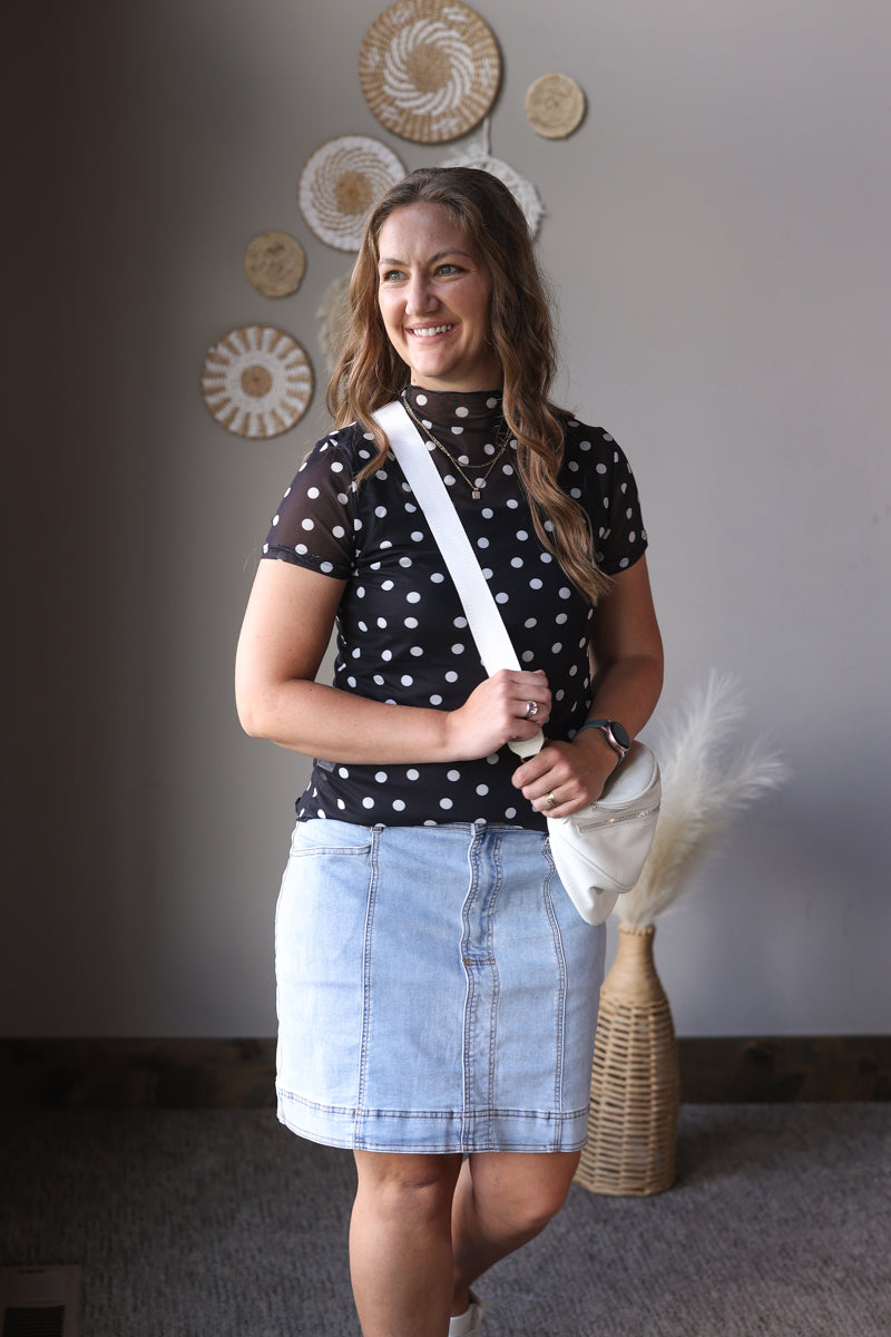 Woman in a polka dot top and denim skirt holding a white bag in a room with decorative wall plates.