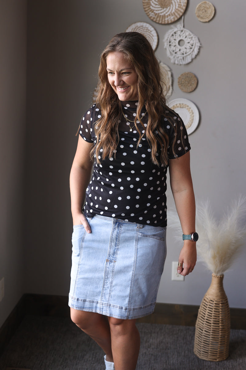 Woman wearing a black polka dot shirt and denim skirt in a room with decorative plates on the wall.