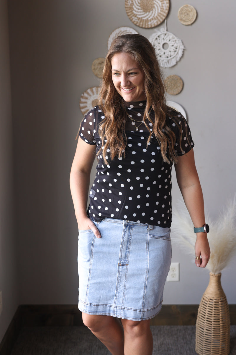 Woman wearing a black polka dot shirt and denim skirt indoors.
