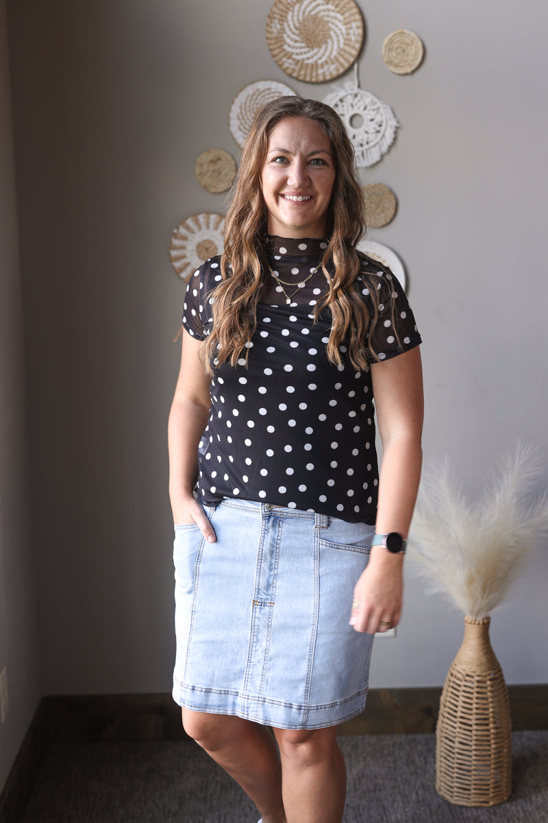Woman wearing a black polka dot top and denim skirt standing against a gray wall with decorative elements.