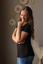Woman wearing a black t-shirt and blue jean skirt standing against a neutral wall with decorative elements.