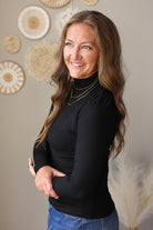 A woman with long brown hair smiling and posing in a black long-sleeve mock neck turtleneck top and blue jeans, standing in front of a beige wall decorated with circular woven wall art.