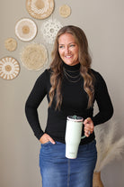 A woman with long brown hair stands indoors holding a pale mint green insulated tumbler with a lid. She is wearing a black long-sleeve top and a blue denim skirt.