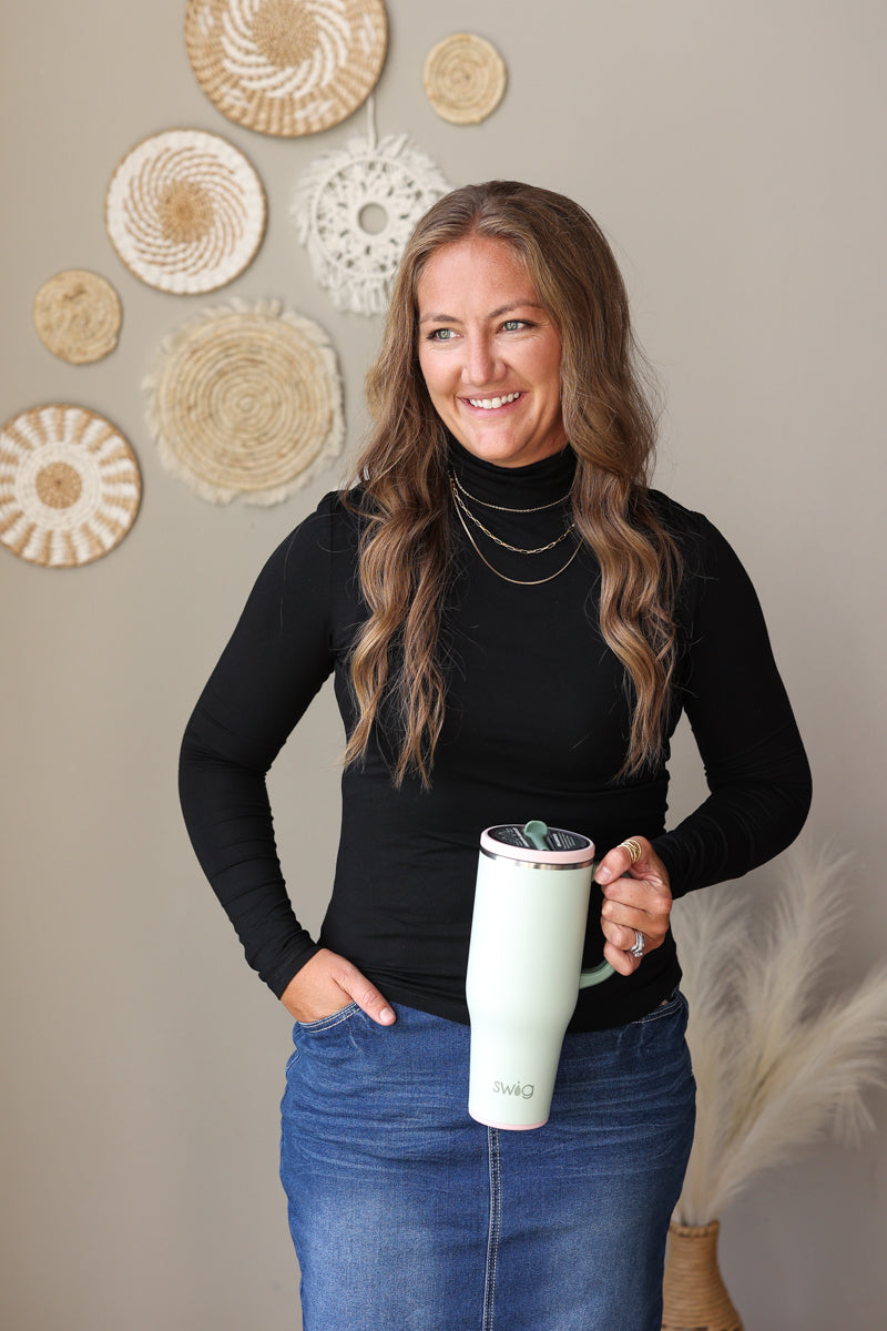 A woman with long brown hair stands indoors holding a pale mint green insulated tumbler with a lid. She is wearing a black long-sleeve top and a blue denim skirt.