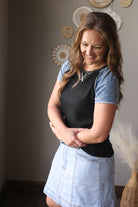 Woman wearing a black top and light blue denim skirt with arms crossed, standing against a neutral wall.