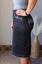 A person wearing a fitted black denim pencil skirt with contrast white stitching and decorative pocket detailing, standing indoors against a neutral wall.