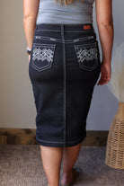 A person shown from the back wearing a dark denim pencil skirt with white contrast stitching and geometric embroidery on the back pockets, paired with a gray top.