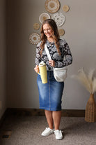 Woman holding a yellow cup and white bag in a room with decorative, wearing a black floral mesh top and midi denim skirt