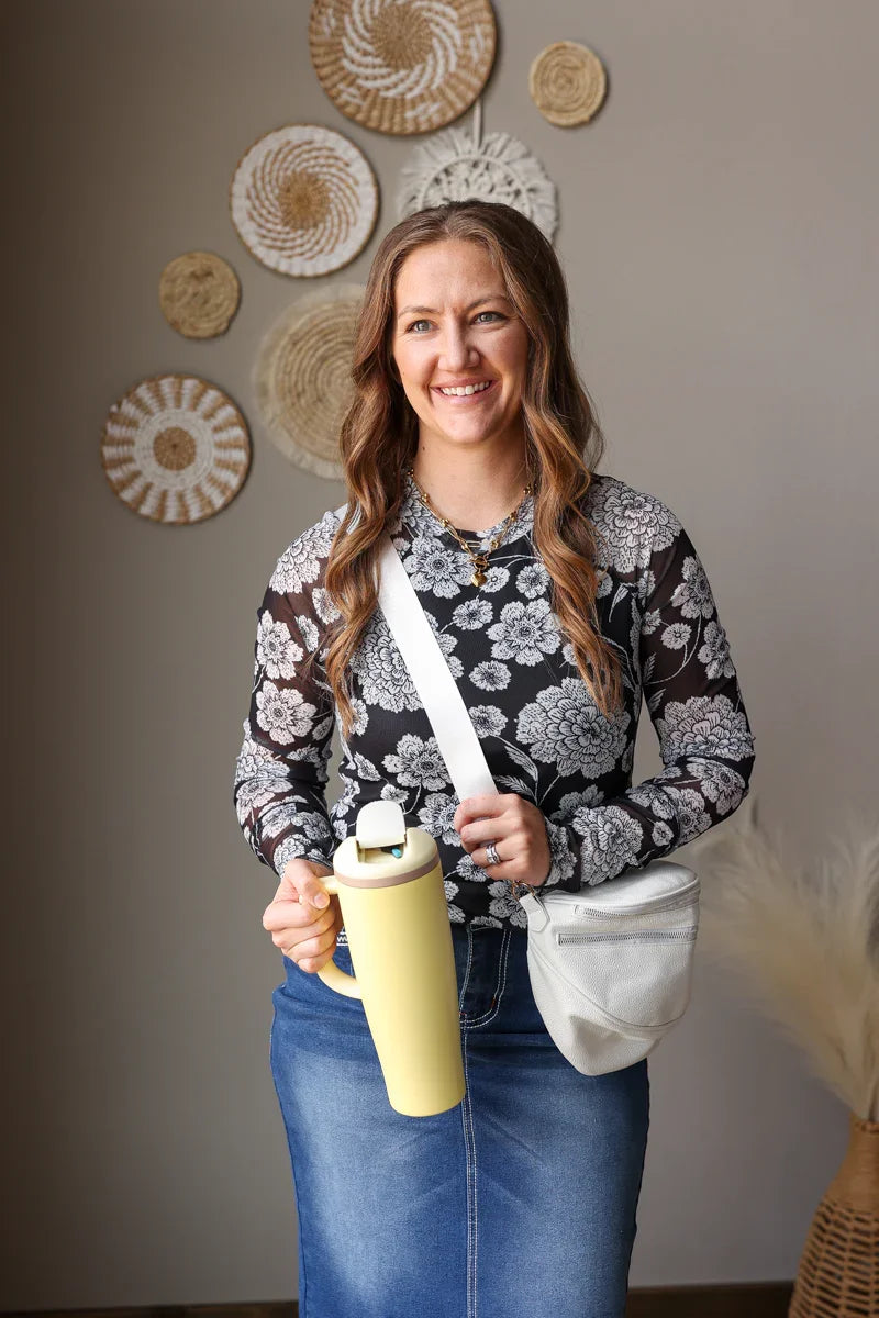 Woman holding a yellow water bottle with decorative plates on the wall behind her, wearing a black white floral mesh long sleeve top for spring. 