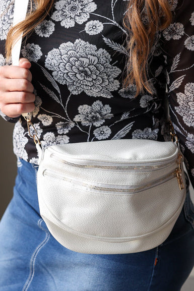 Woman wearing a black floral top and blue skirt holding a white fanny pack crossbody purse. 
