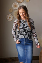 Woman wearing a floral long-sleeve top and blue skirt standing against a neutral wall with decorative items.