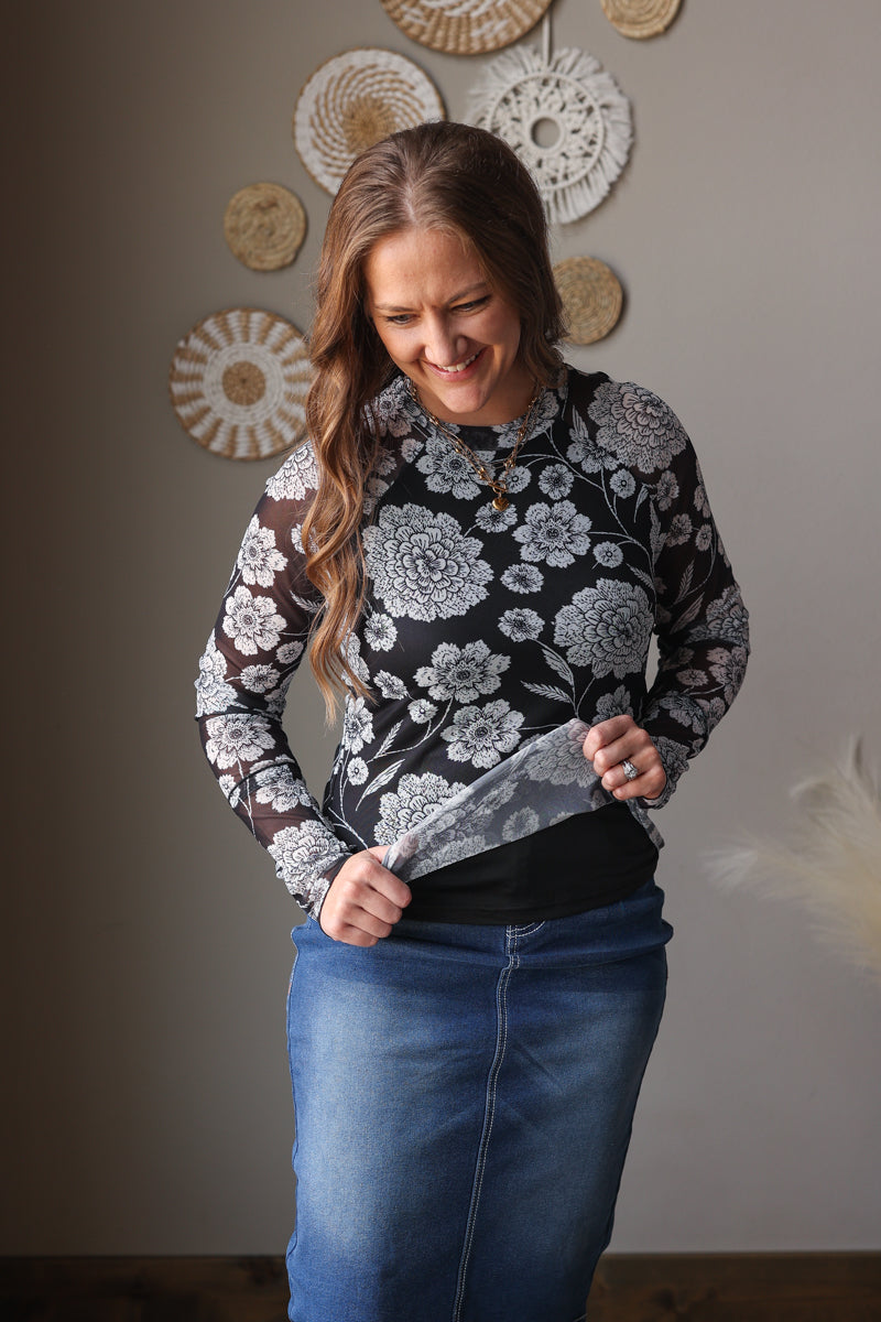 Woman wearing a floral mesh top and blue jean skirt standing against a neutral wall with decorative plates.