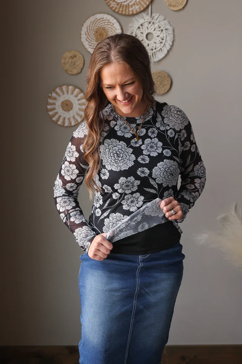 Woman wearing a floral mesh top and blue jean skirt standing against a neutral wall with decorative plates.