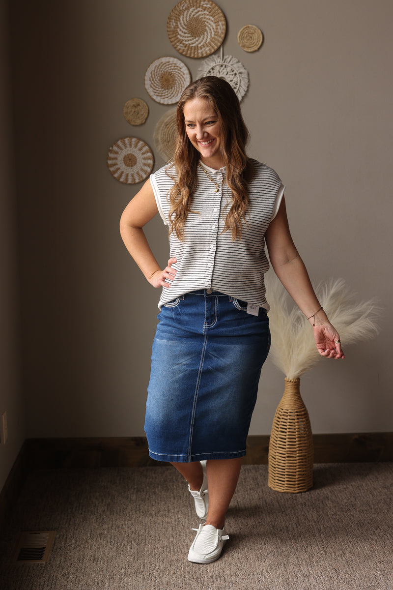 Woman in a striped shirt and denim skirt holding a decorative fan in a room with wall art.