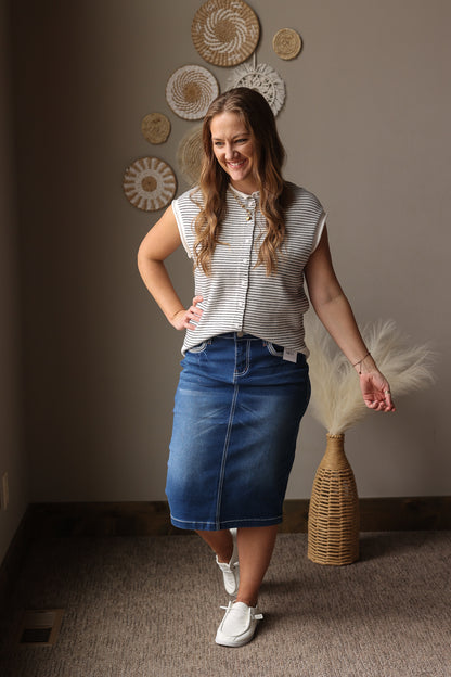 Woman in a striped shirt and denim skirt holding a decorative fan in a room with wall art.