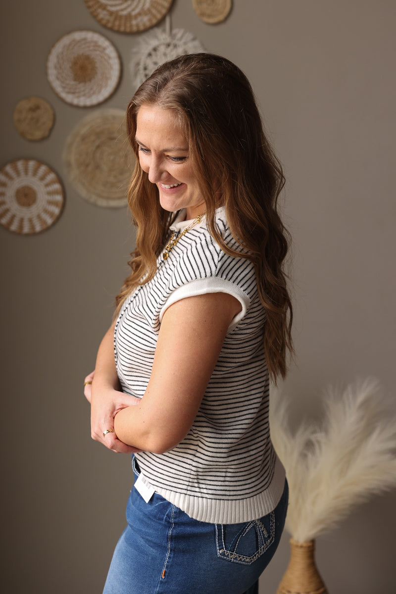 Woman wearing a striped shirt and blue jeans standing against a wall with decorative items.