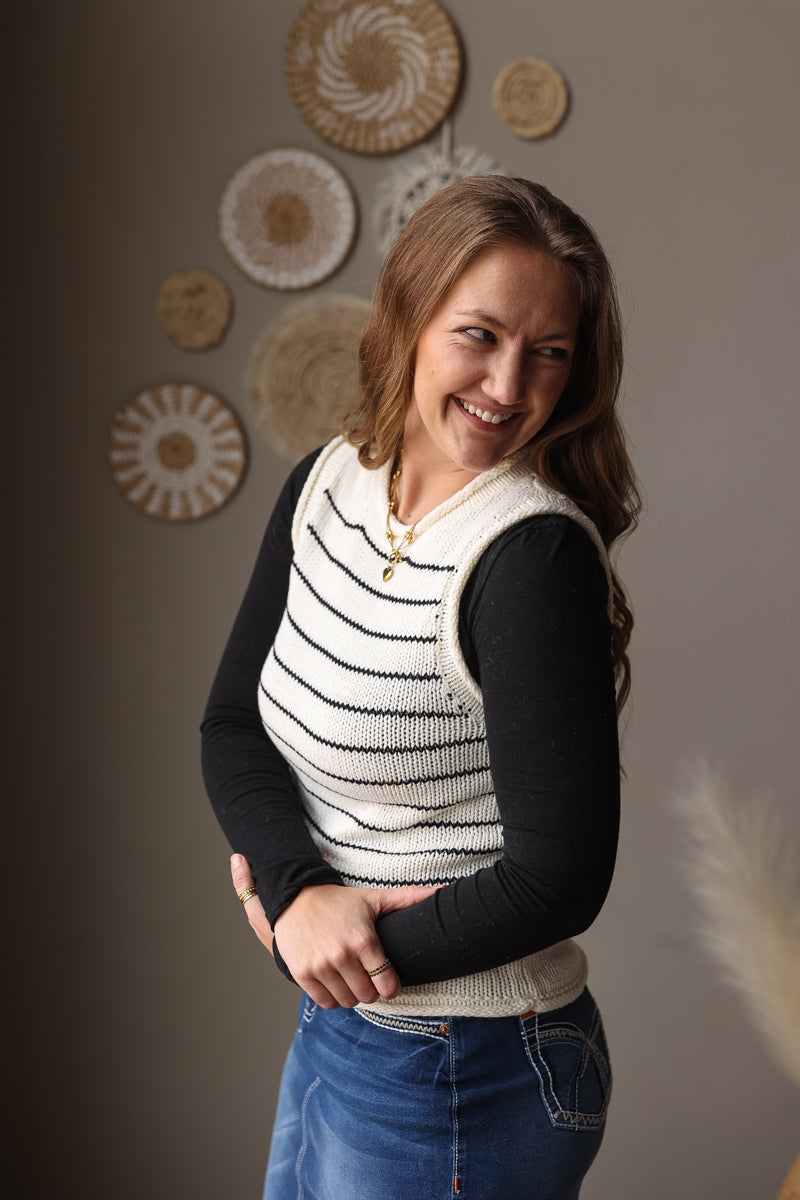 Woman wearing a black and white striped vest over a black long-sleeve shirt, standing against a wall with decorative plates.