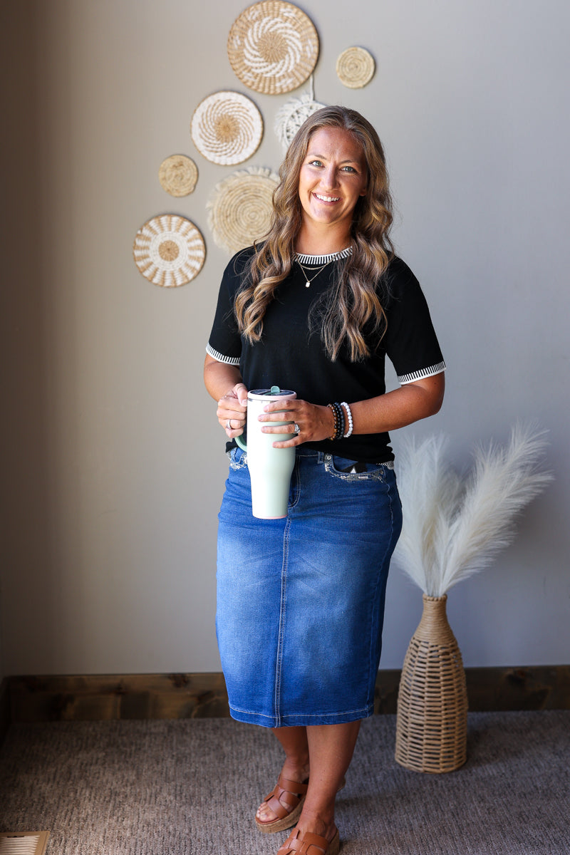 A woman stands indoors wearing a black short-sleeve top with white trim and a blue denim skirt, holding a white tumbler. The background shows a gray wall with decorative woven wall art and a woven vase with pampas grass.