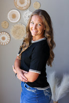 A smiling woman with long wavy brown hair stands indoors wearing a black short-sleeve top with white striped trim at the collar and cuffs, paired with blue jean skirt. The background is a gray wall decorated with circular woven wall art and white pampas grass.