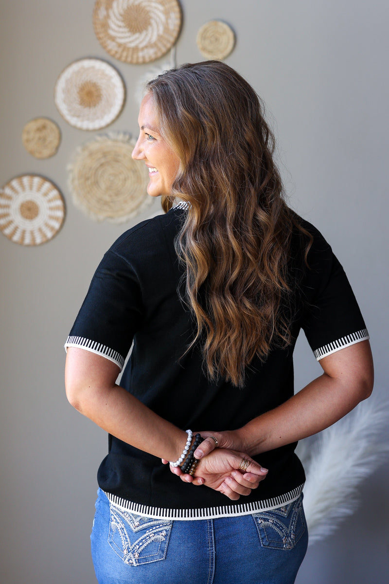 A woman with long wavy brown hair is shown from the back, wearing a black short-sleeve top with white striped trim on the sleeves and hem, paired with blue jeans. She is standing against a gray wall decorated with circular woven wall art.