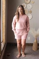 Woman wearing a pink textured sweatshirt and shorts outfit standing in a room with decorative plates on the wall.
