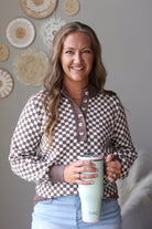 A smiling woman with long light brown hair stands indoors holding a mint green insulated tumbler with a lid. She wears a brown and beige checkered long-sleeve henley-style top and light blue jean skirt.