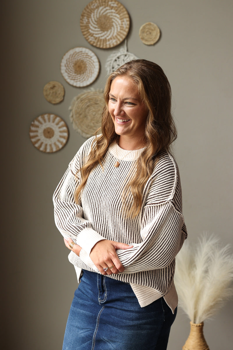 A woman smiling and looking to the side, wearing a cream and dark brown vertically striped knit sweater and blue jean skirt, standing in front of a neutral wall decorated with woven wall art.
