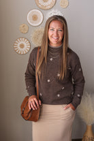 A smiling woman stands indoors wearing a brown knit sweater with small white embroidered star-like motifs, a beige skirt, layered silver necklaces, and a brown crossbody bag. The background shows a neutral wall with decorative woven wall art and a vase with dried grass.
