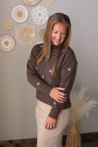 A woman wearing a brown knit sweater with small white embroidered floral details, paired with beige skirt, standing in a decorated room with wall art and a vase of pampas grass.