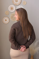 A woman with long brown hair is shown from the back wearing a dark brown knit sweater and a beige skirt, standing in front of a neutral wall decorated with circular woven wall art.